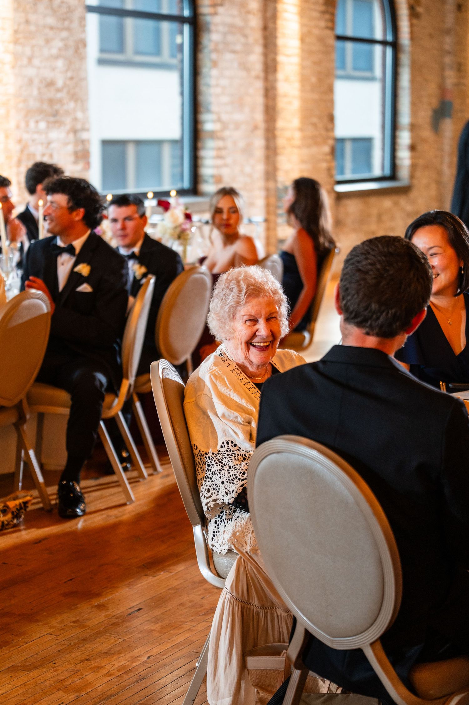 Elderly woman laughing joyfully at a wedding reception in a brick-walled venue, surrounded by elegantly dressed guests.