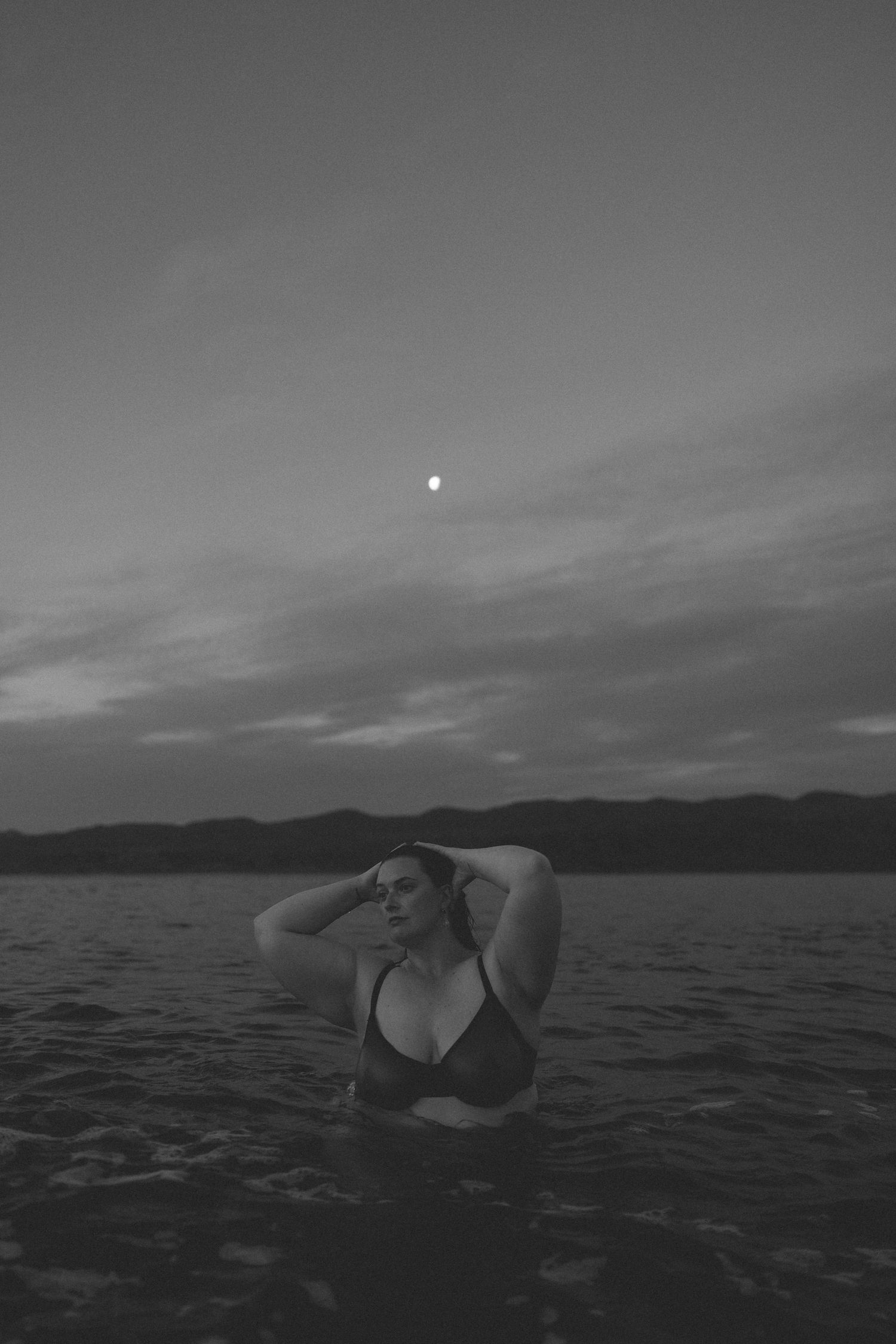 Black and white photo of a woman in a bikini standing in ocean water at dusk with a full moon overhead.