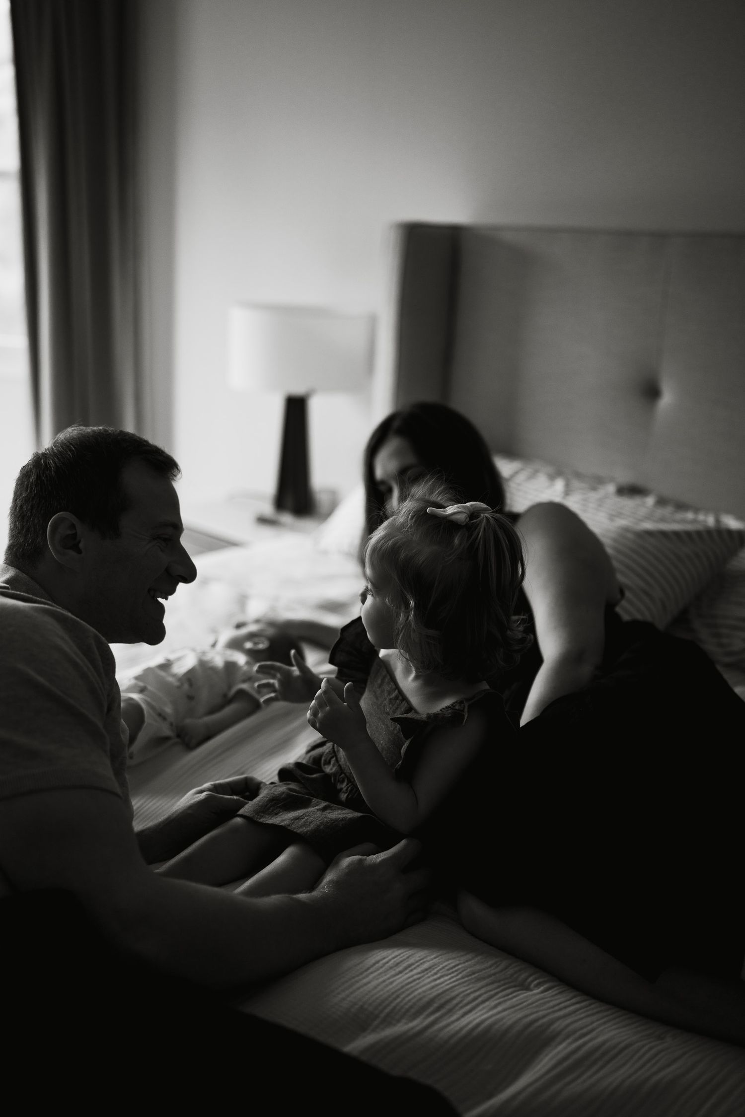 Black and white photo of a family with a young child sharing a tender moment together on a bed in a cozy bedroom.