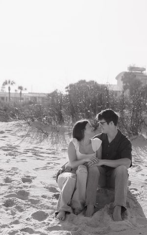 A romantic black and white photo of a couple sitting close together on a sandy beach with palm trees in the background.