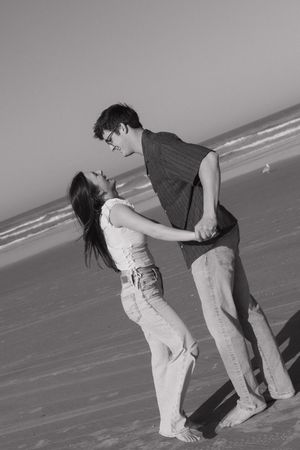 A couple playfully dances together on a beach in black and white, creating a romantic silhouette against the ocean.