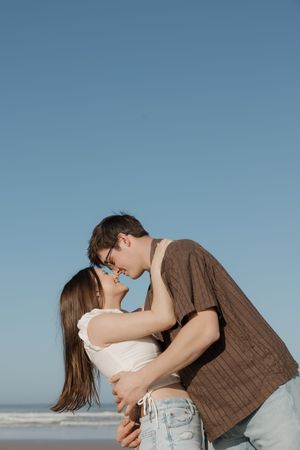 A romantic couple embraces and kisses on a beach against a blue sky background.