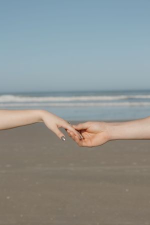 Two hands reaching out to touch each other gently on a peaceful beach with ocean waves in the background.