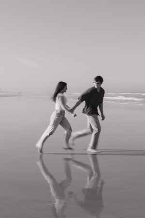 Two people playfully chase each other on a misty beach, creating reflections in the wet sand as waves roll in the distance.