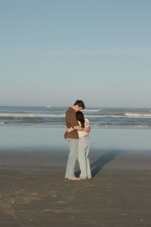 A romantic couple embraces on a serene beach with gentle waves rolling in the background.