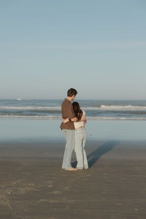 A couple stands together on a serene beach looking out at the ocean waves during golden hour.