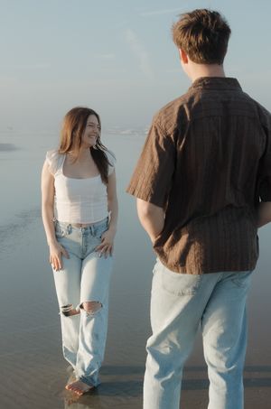 A couple in casual attire stands together on a beach looking out at the ocean during sunset.