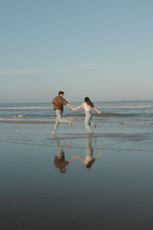 Two people playfully dance and create reflections in the wet sand at the beach during sunset.