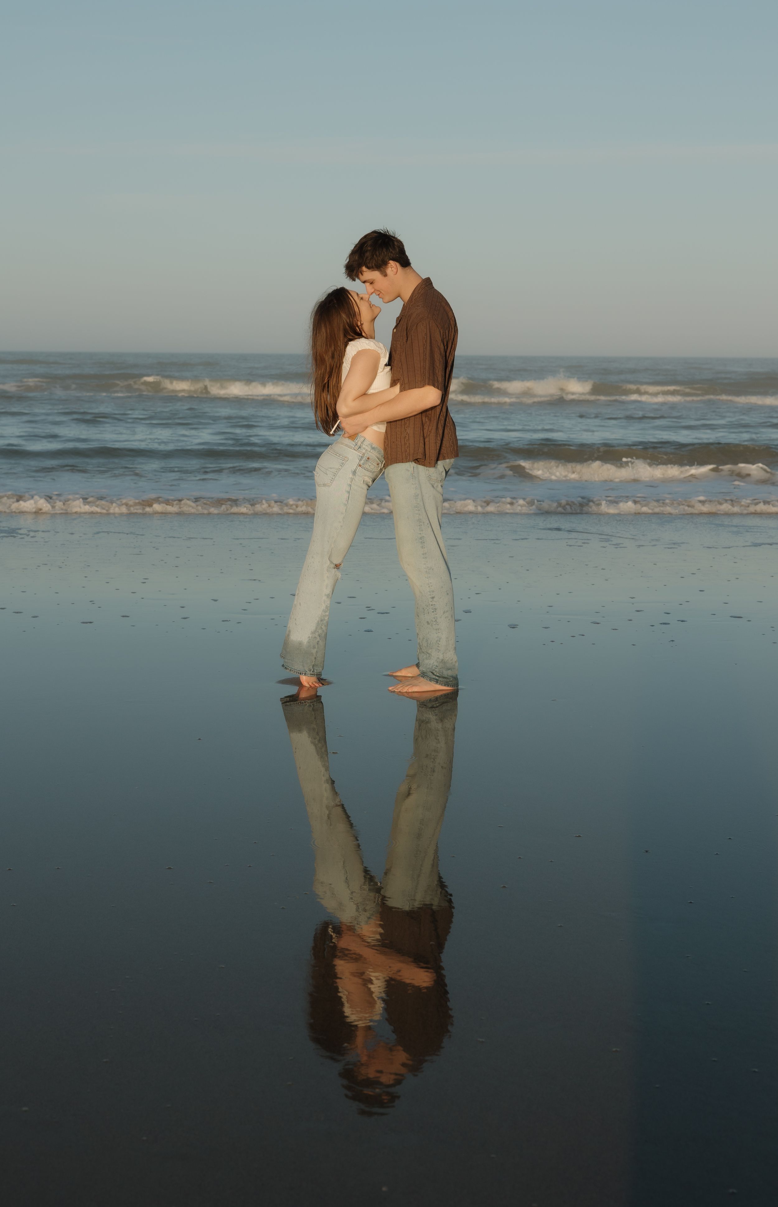 Two people share an intimate moment on a reflective beach at dusk with ocean waves behind them.