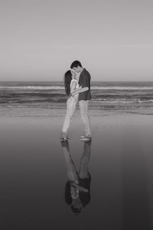 Two people kiss on a reflective beach shore with their mirror image visible in the wet sand.