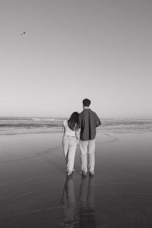 A black and white photo of two people standing together on a beach looking out at the ocean.