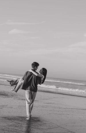 Black and white photo of a couple playing on the beach with one person carrying the other near the waves.