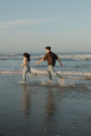 Three people playfully splash and kick through shallow ocean waves at the beach during sunset.