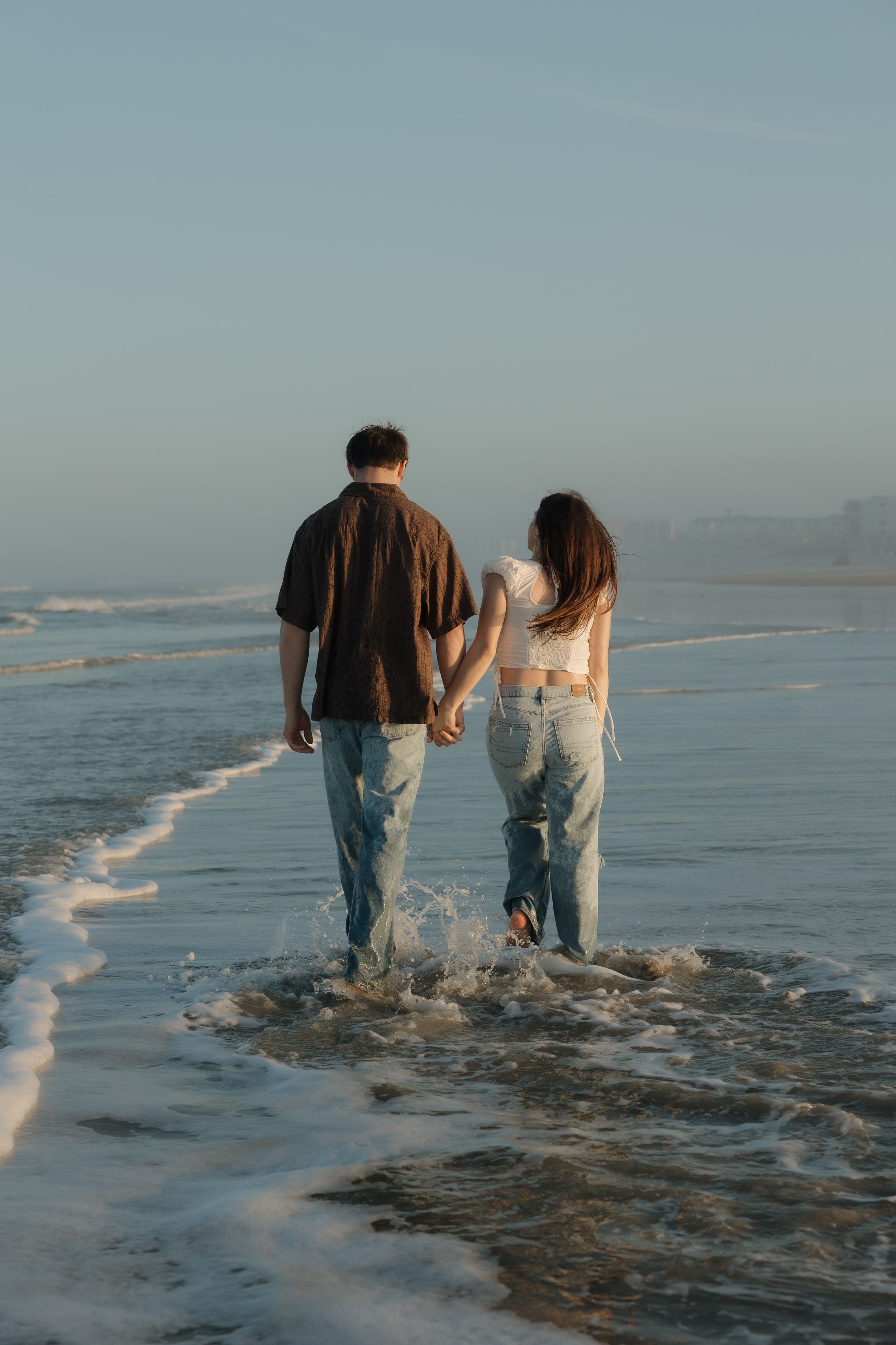 Two people in matching jeans walk barefoot along a misty beach shoreline at sunset.
