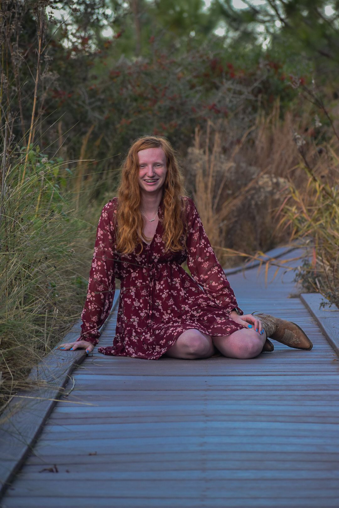 Young redhead woman in floral burgundy dress sitting cross-legged on wooden boardwalk path surrounded by tall grasses.