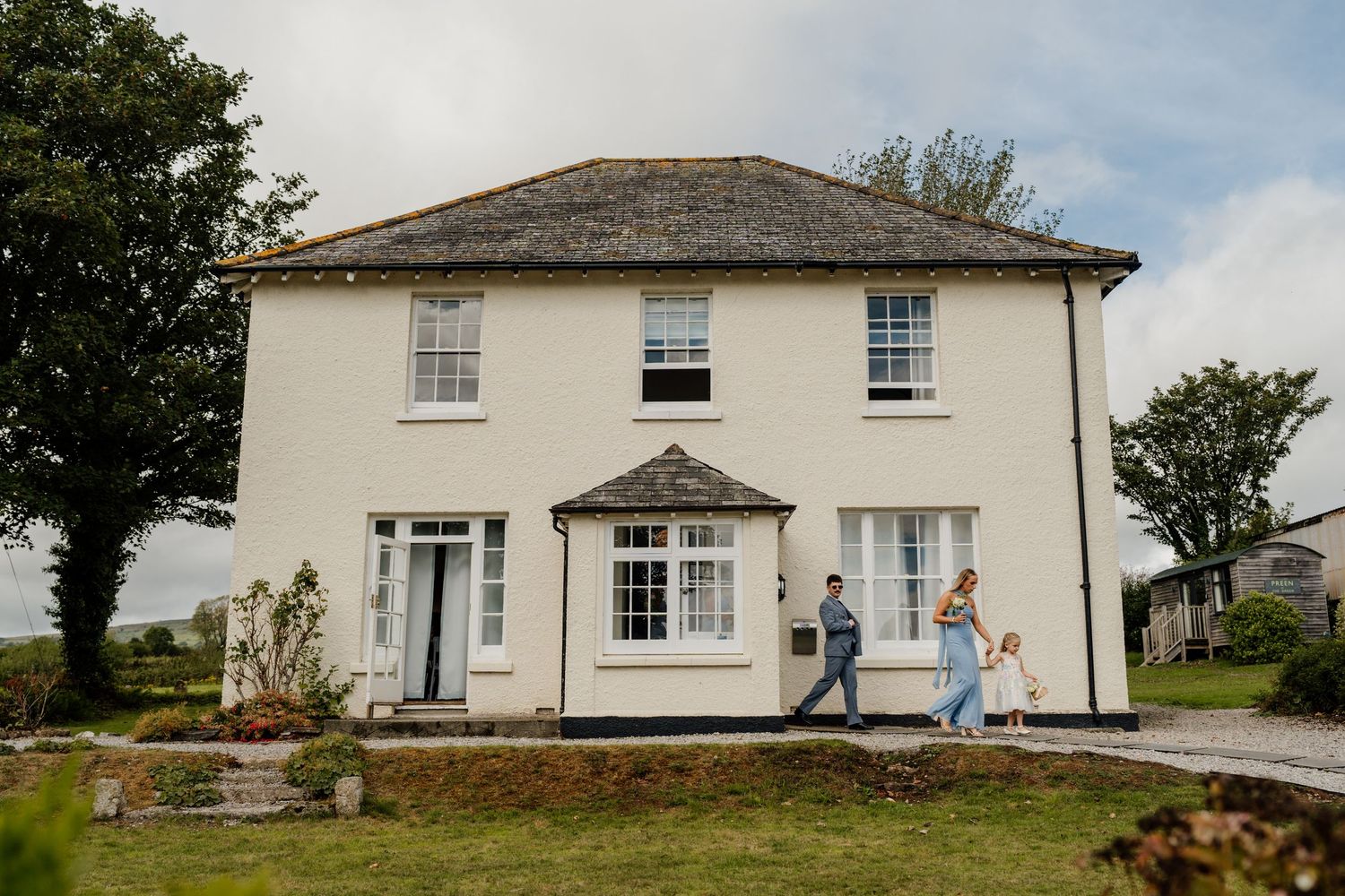 Wedding party walking outside a charming white cottage with lush green gardens at The Green wedding venue in Liskeard.