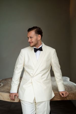 A man in a cream white tuxedo with black bow tie leans against a wooden table, looking sideways.