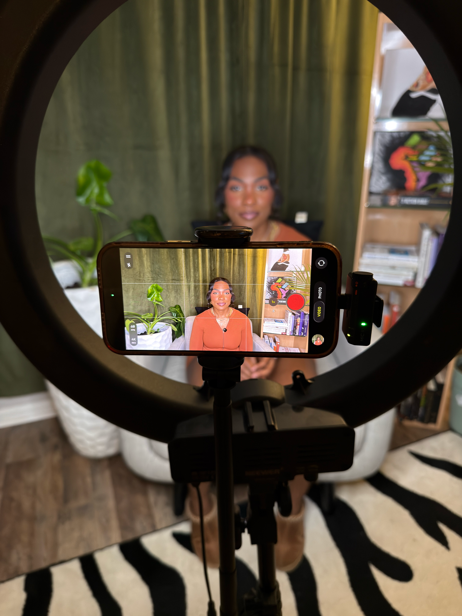 Woman recording herself on a smartphone mounted on a ring light tripod setup in a cozy home studio space.
