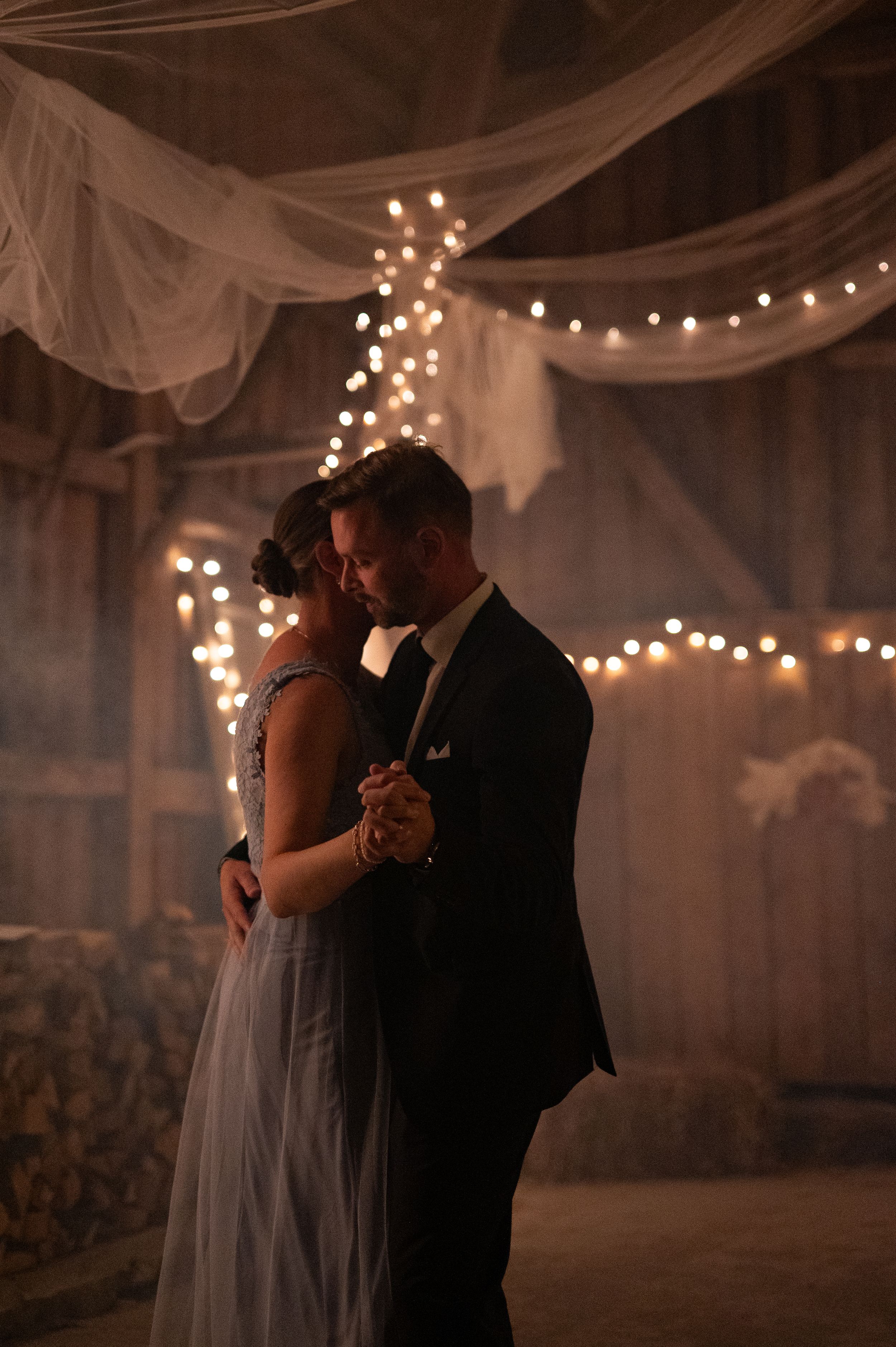 Couple shares first dance in a romantic barn wedding venue adorned with fairy lights and flowing white drapes.