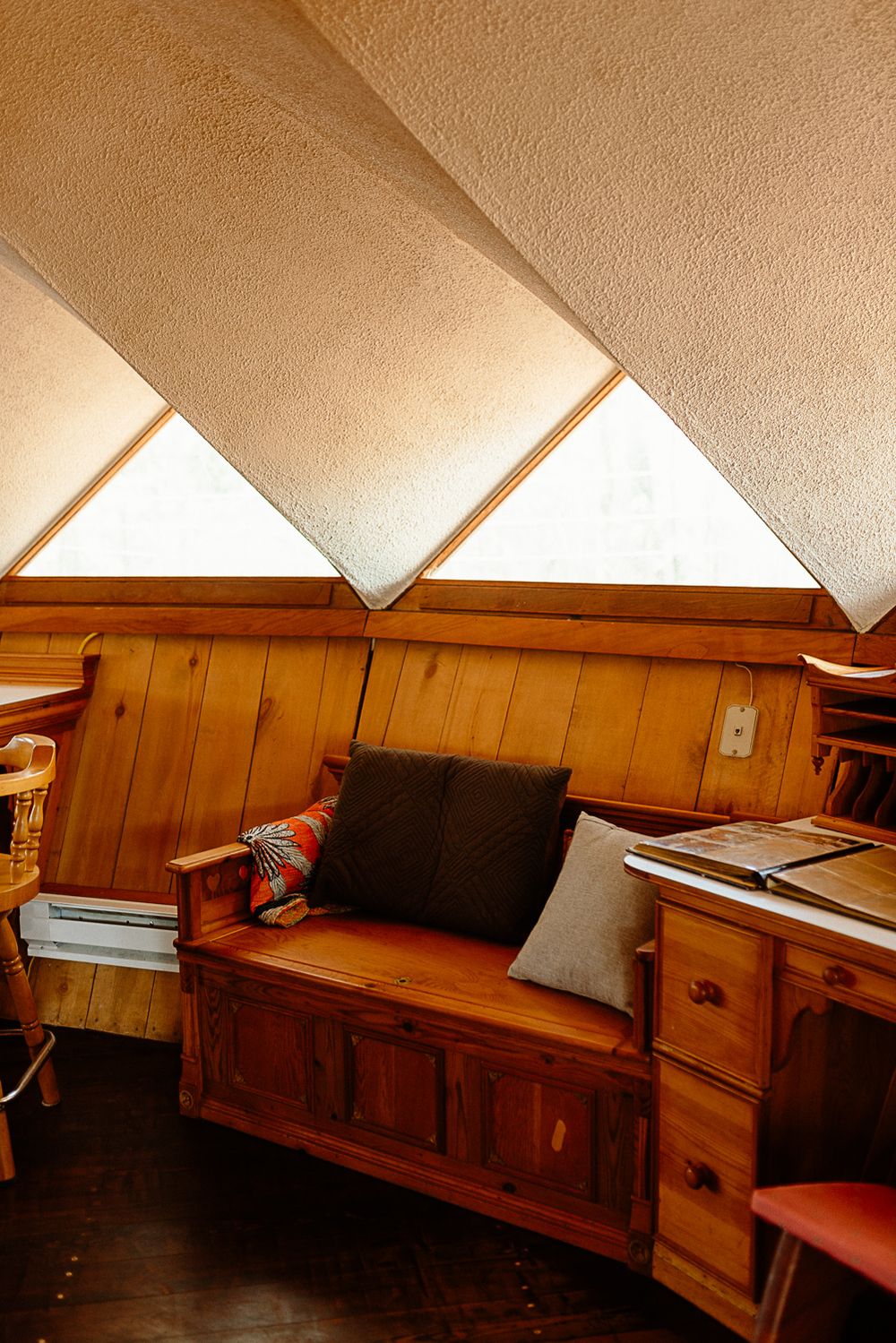 Cozy wooden bench with pillows beneath triangular skylights in a rustic wood-paneled interior nook.