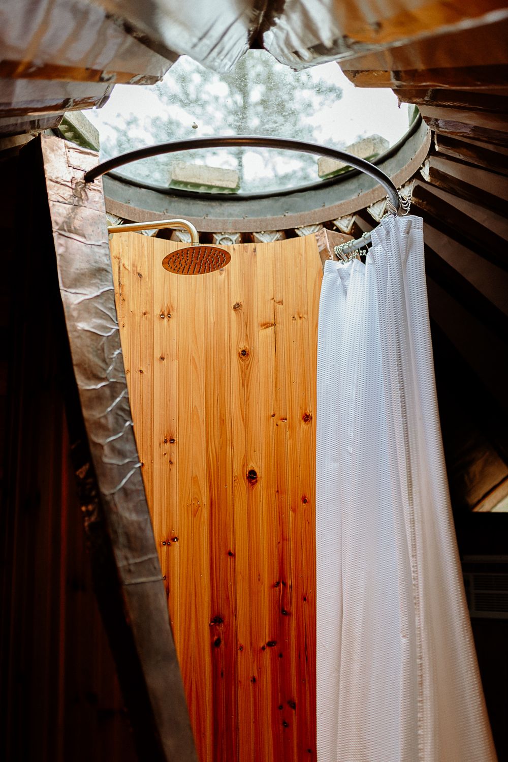 Circular shower with rain head, wooden slat wall, and white curtain inside a rustic cabin bathroom.