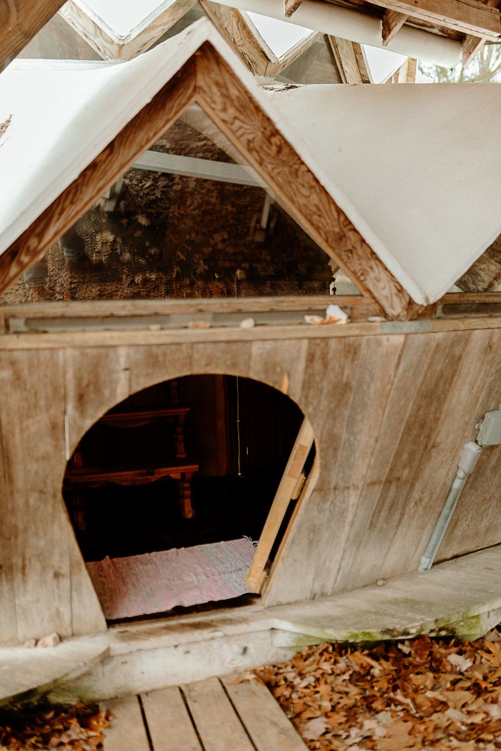 Rustic wooden chicken coop with arched entry door and triangular roof details on a wooden deck surrounded by autumn leaves.
