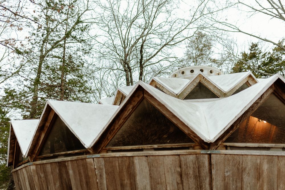 Snow-covered triangular wooden roof peaks of a rustic outdoor structure surrounded by bare winter trees.