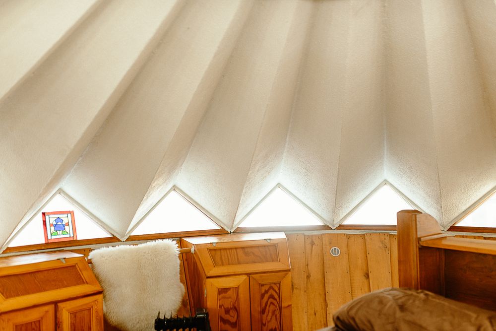 Interior of a rustic yurt with wooden furniture, white canvas ceiling panels, and triangular windows letting in natural light.