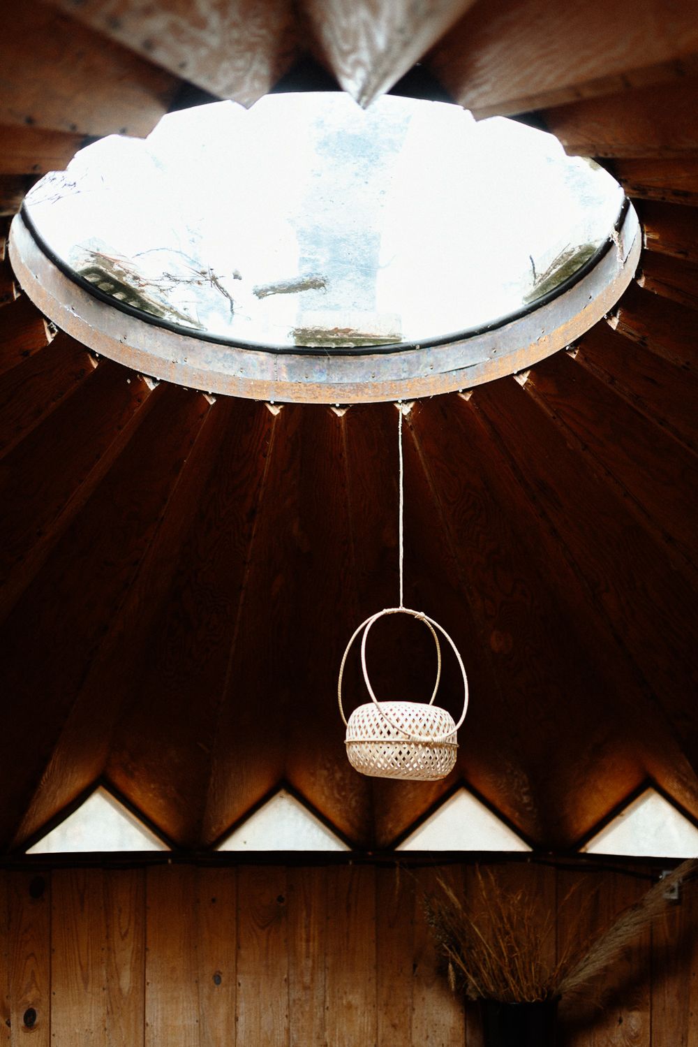 Rustic wooden ceiling with circular skylight and hanging wicker basket lantern in a cozy cabin interior.