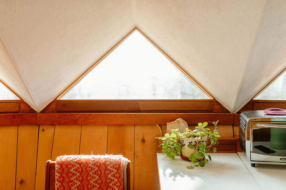 Cozy A-frame cabin interior with triangular windows, wood-paneled walls, a potted plant, and a red patterned chair.