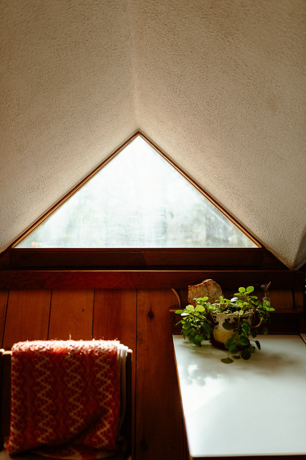 Cozy attic room with triangular window, wooden walls, potted plant on white table, and red blanket on chair.