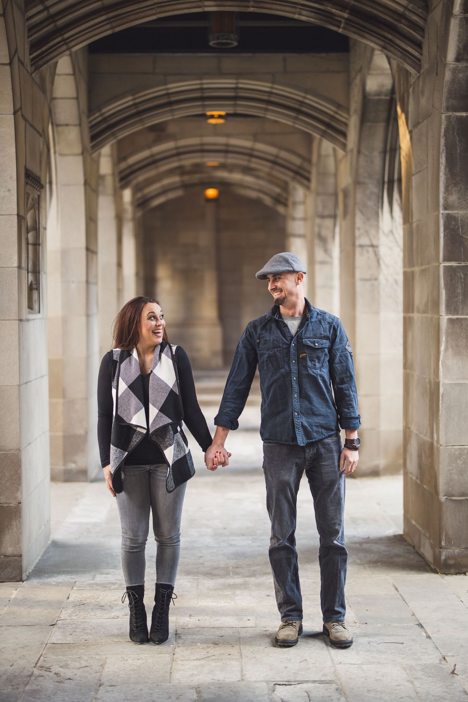 A smiling couple holding hands while walking through a stone archway, she wears a plaid vest and boots, he wears a denim shirt and flat cap.