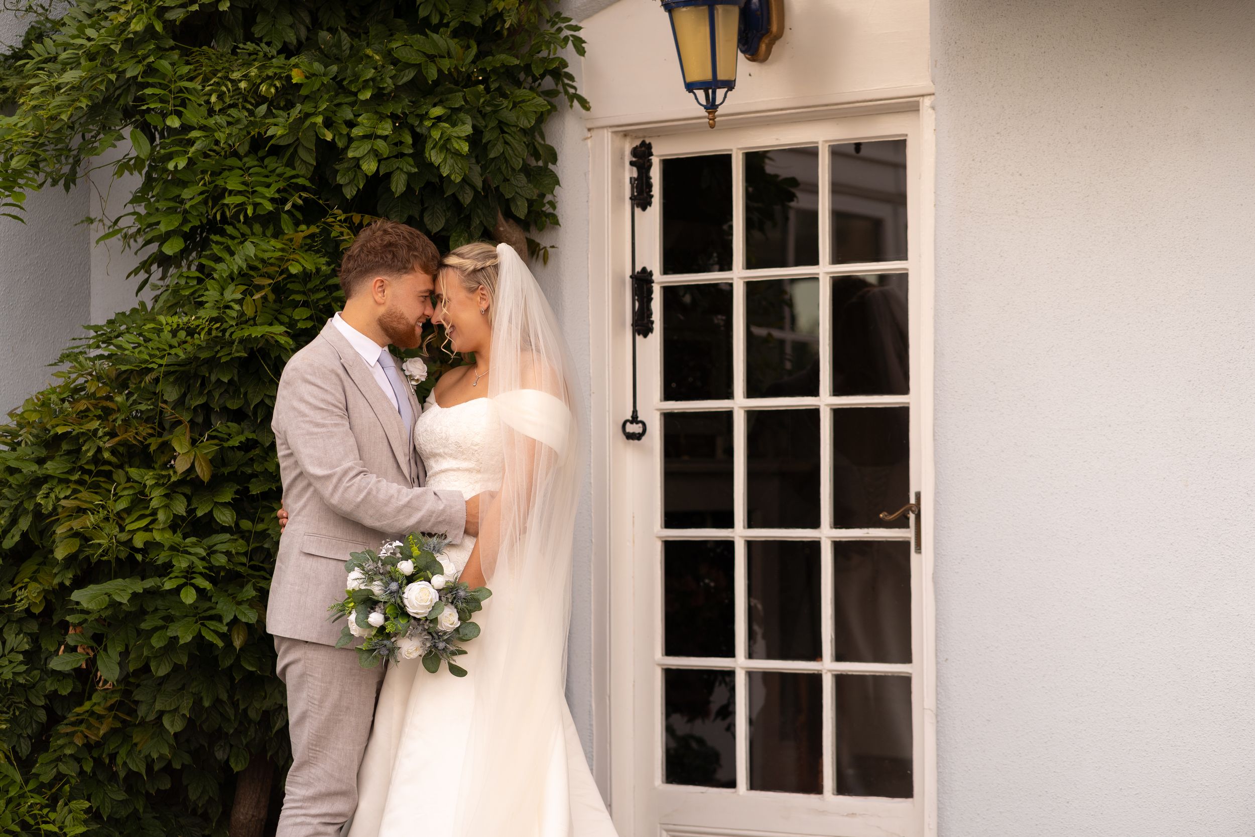 Bride and groom share a romantic moment outside a white building, bride holding a white bouquet, groom in a gray suit.