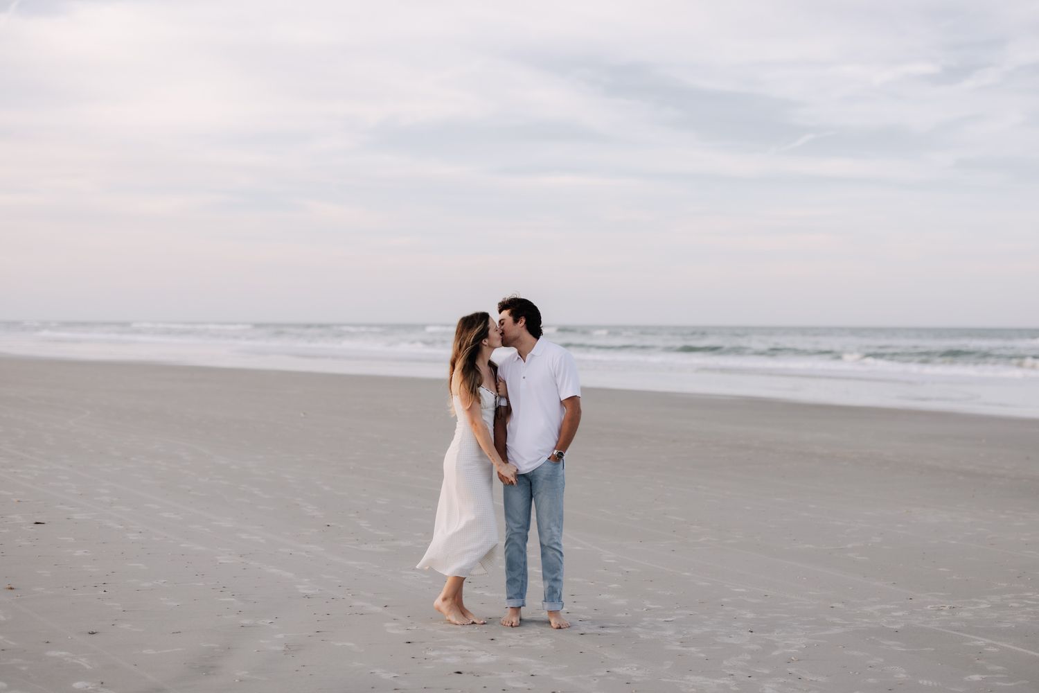 Couple sharing a romantic kiss on a sandy beach with ocean waves and cloudy sky in the background.