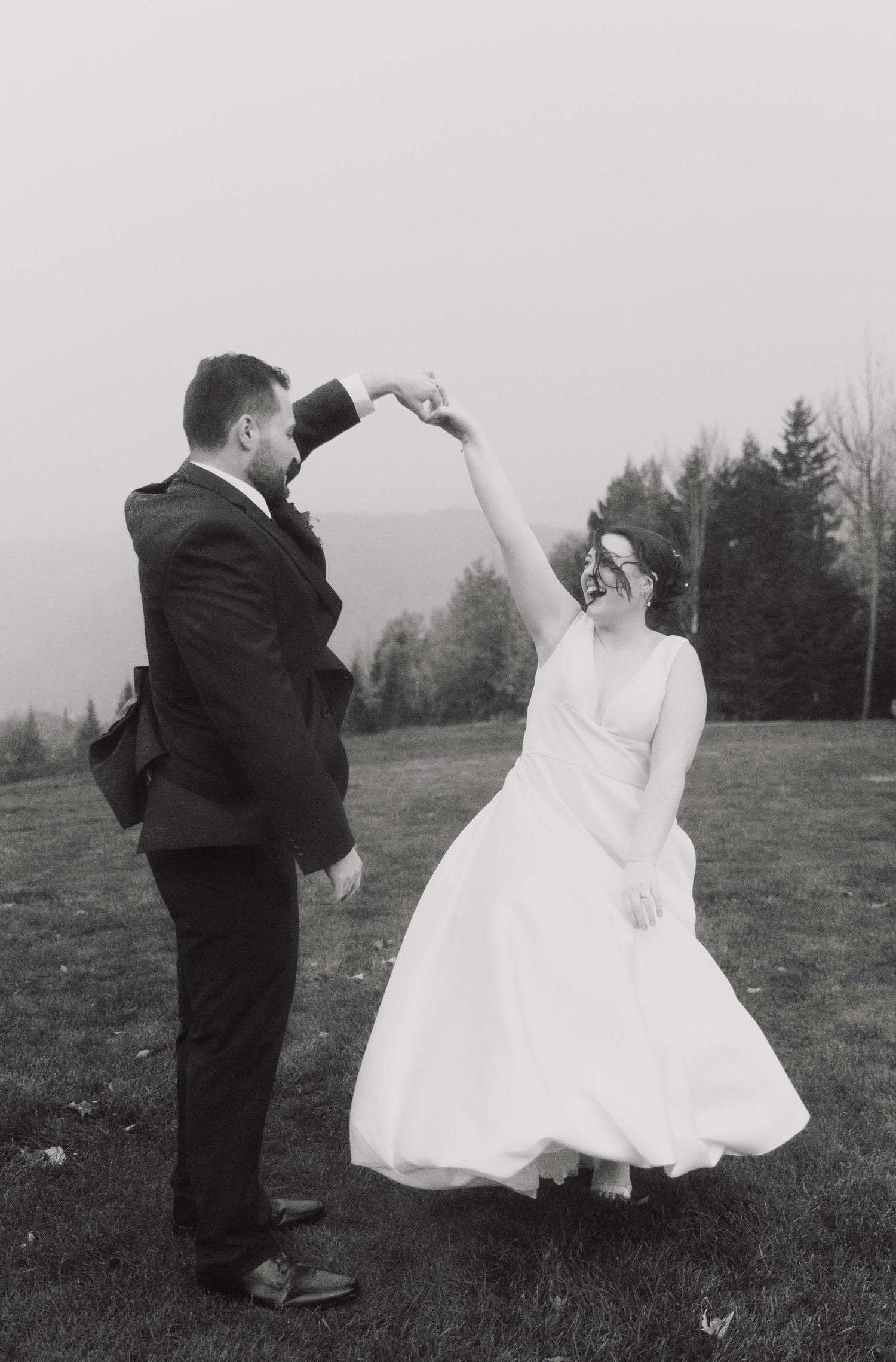 Black and white photo of a joyful bride and groom dancing outdoors, groom spinning bride in wedding dress on misty day.