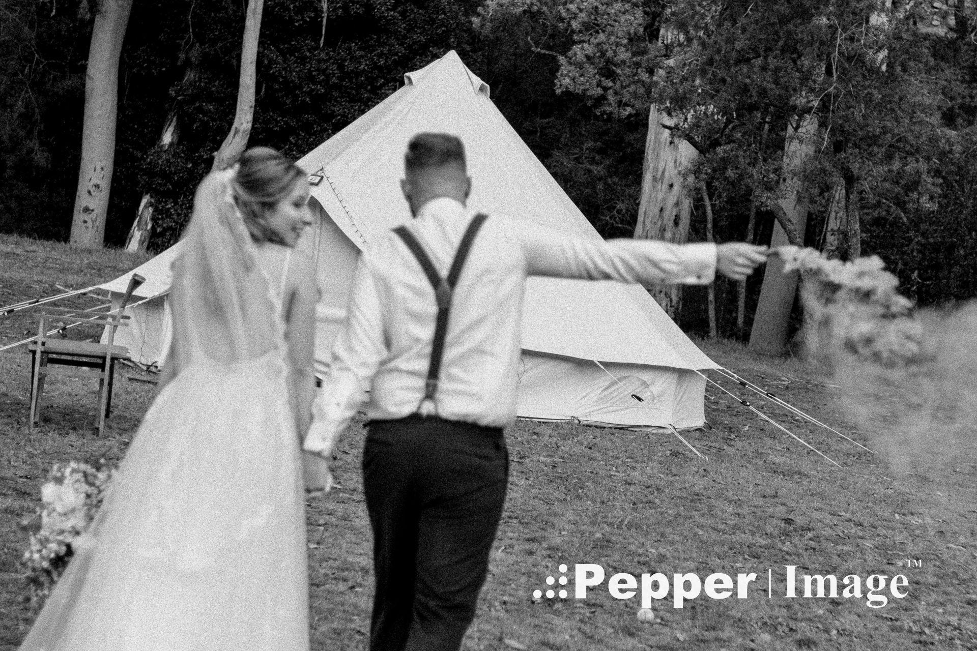 Black and white photo of bride and groom walking toward bell tent with smoke flare at outdoor wedding.