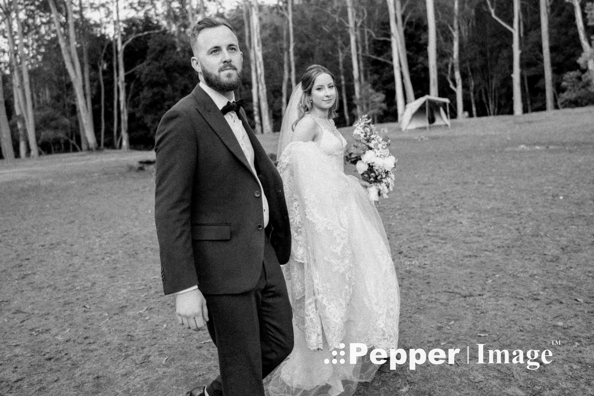 Black and white portrait of bride and groom walking through forest clearing on wedding day.