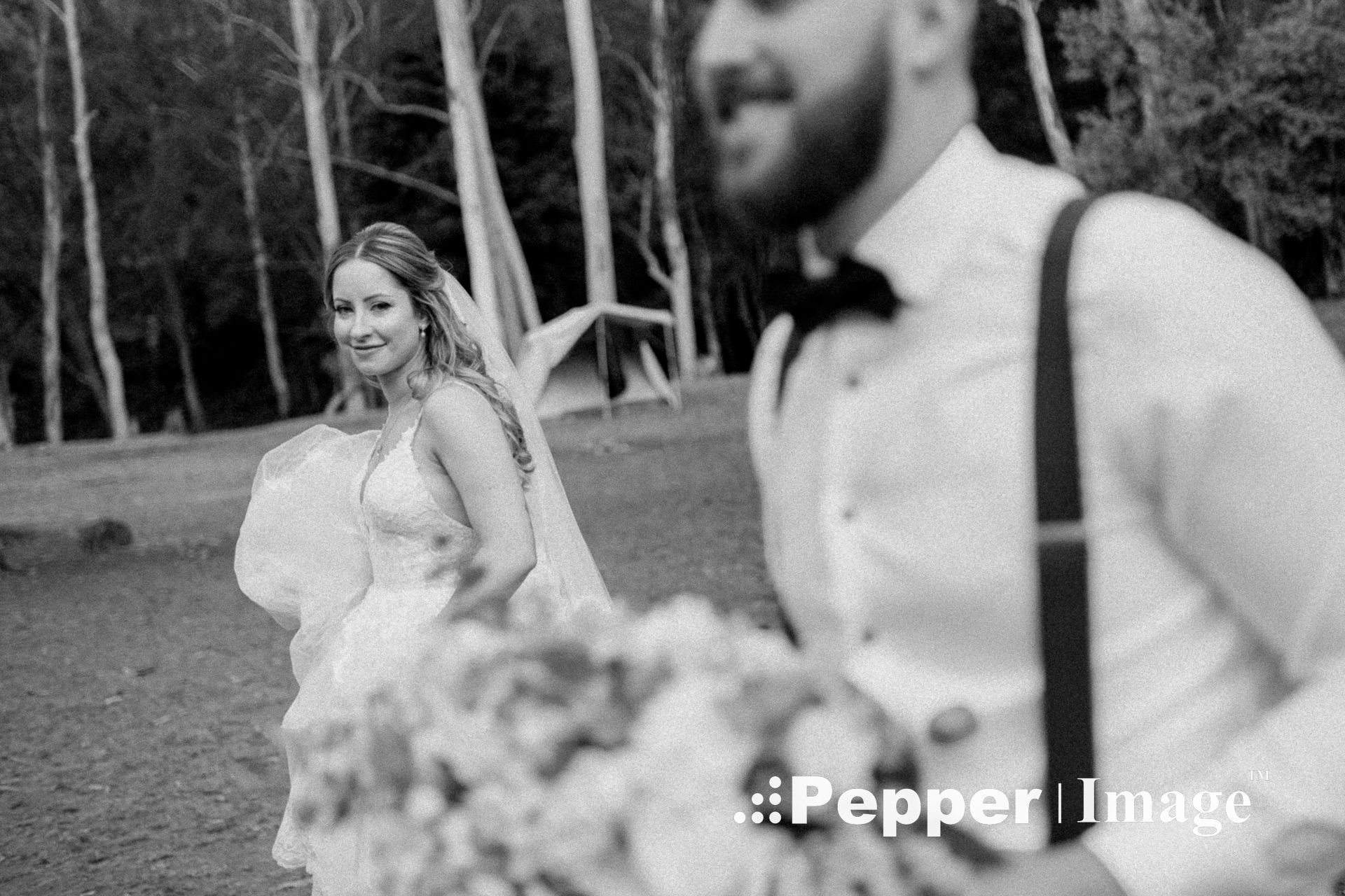 Black and white close-up of smiling bride holding bouquet with groom in suspenders in foreground.