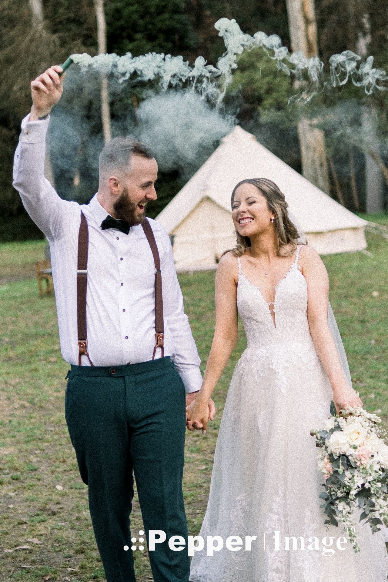 Groom holds green smoke flare while holding hands with bride in lace gown at boho outdoor wedding.