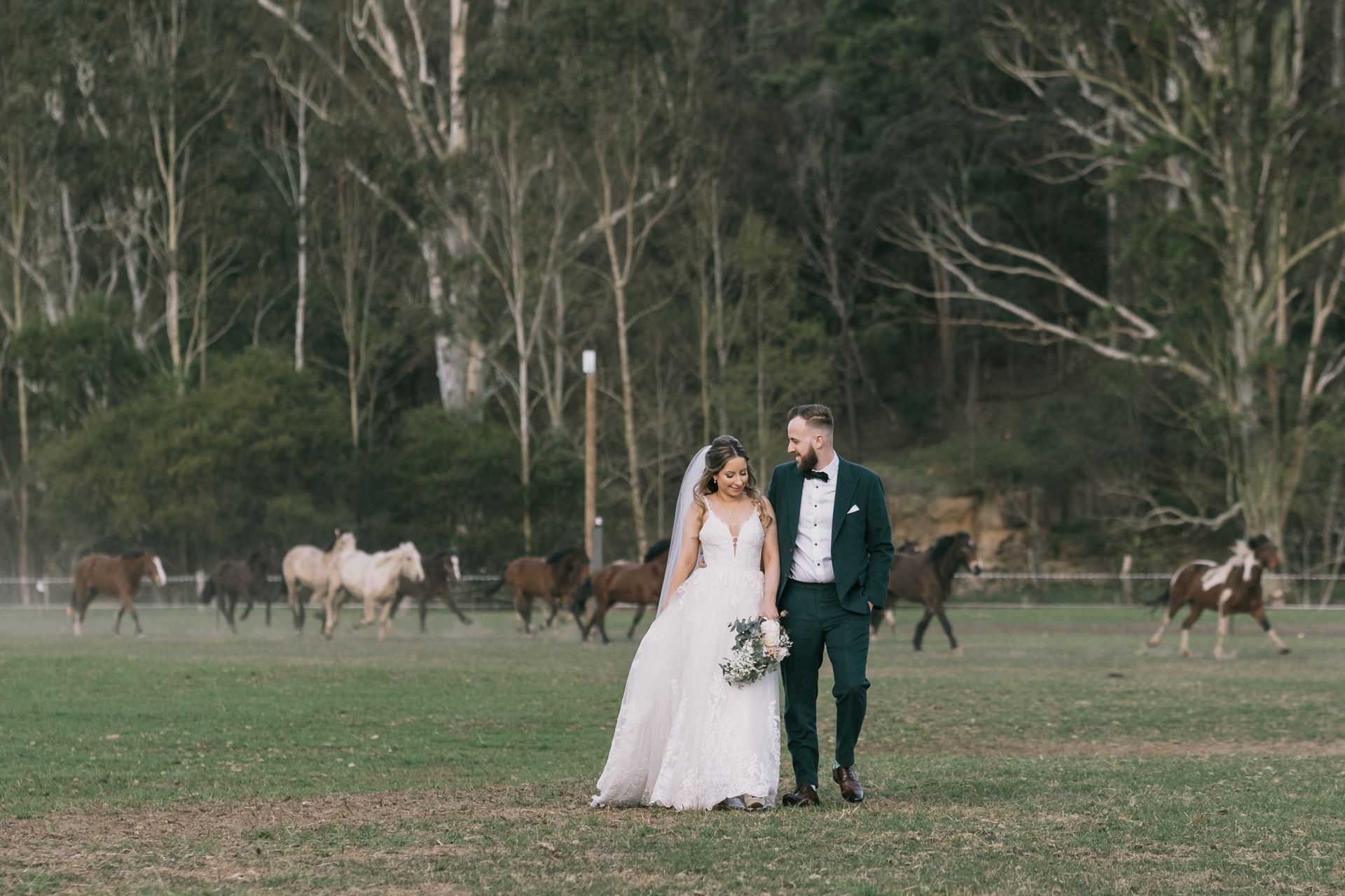 Bride and groom share romantic moment in open field with horses grazing in the background.