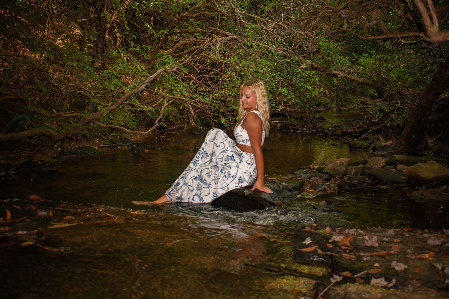 Blonde woman in a floral maxi dress sitting gracefully on rocks in a peaceful woodland stream.
