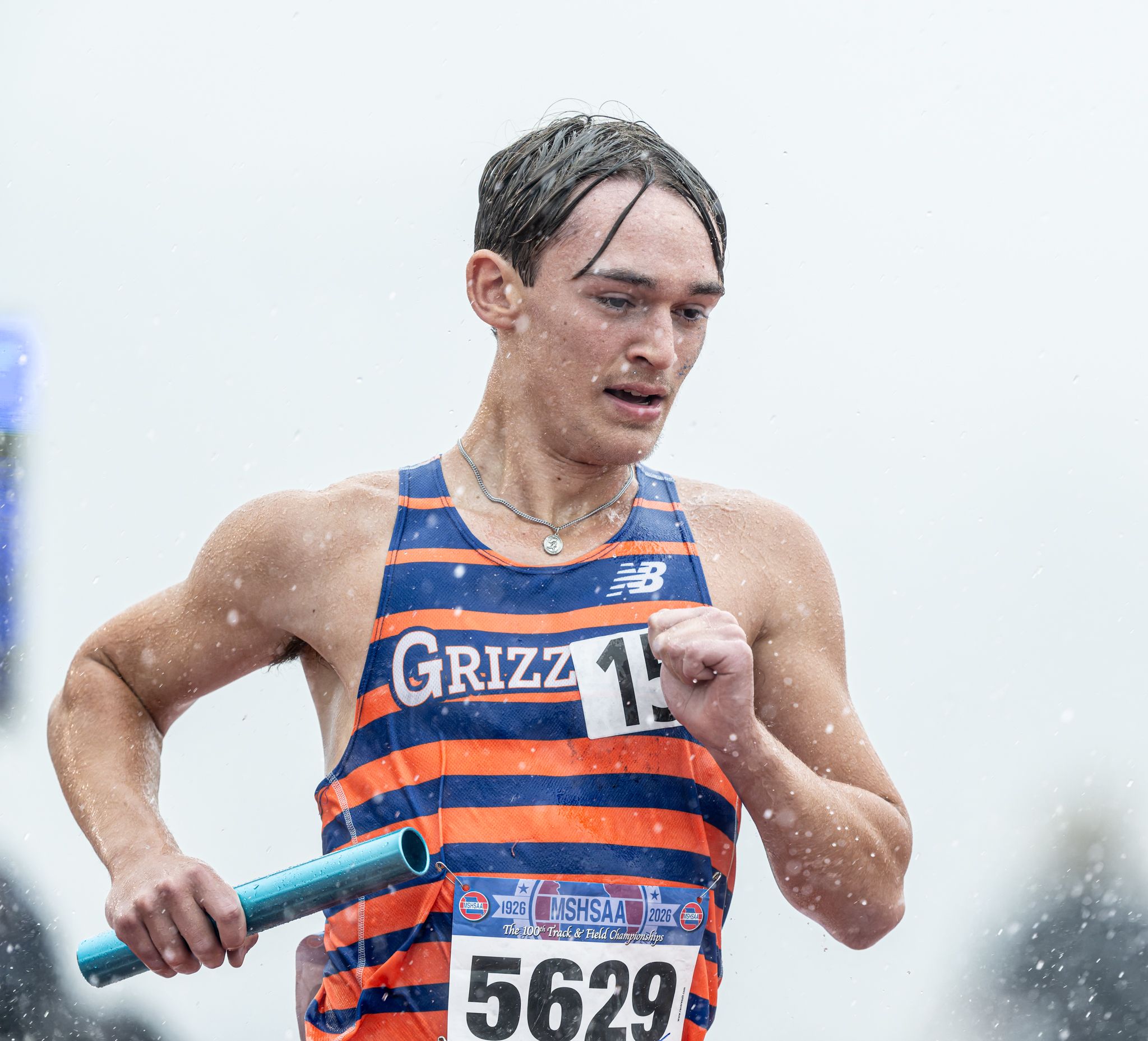 Male runner in Grizzly uniform, number 5629, races in rainy conditions holding a blue baton.
