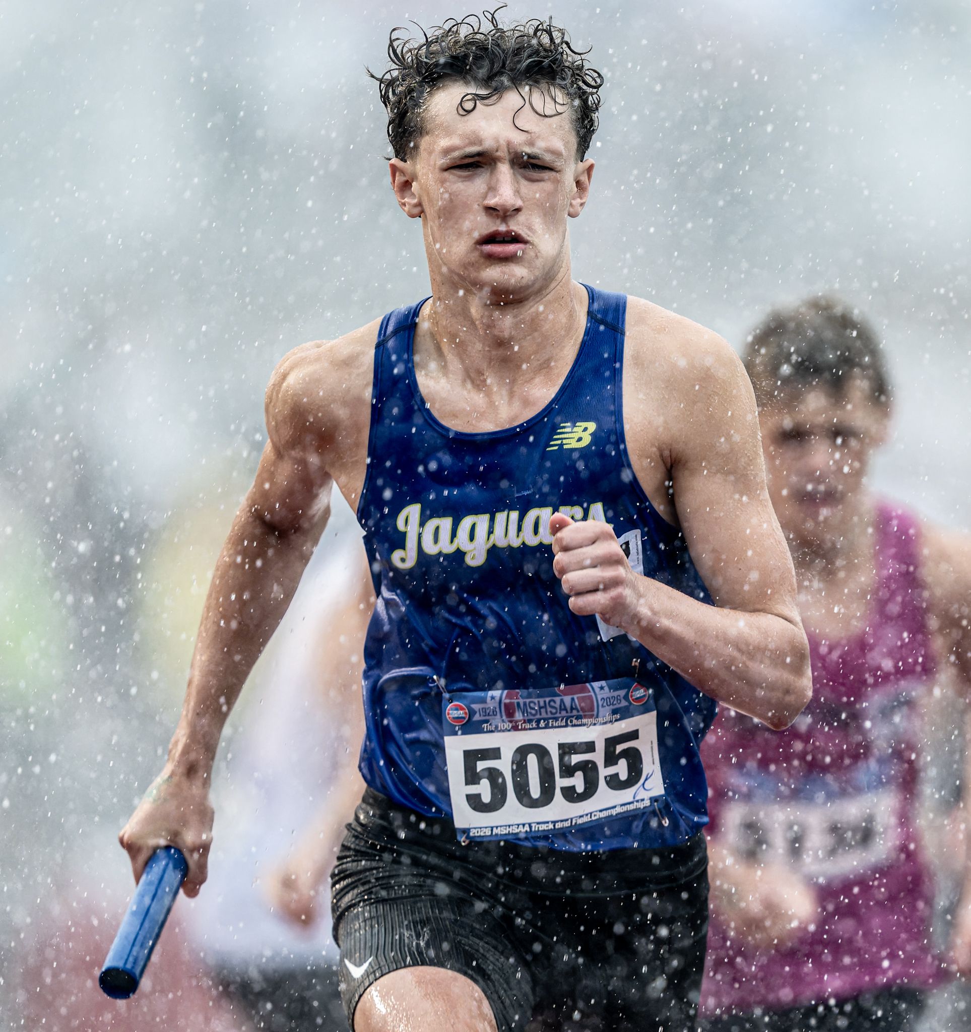 Runner wearing Jaguars jersey number 5055 competes in a relay race during heavy rain at a track meet.