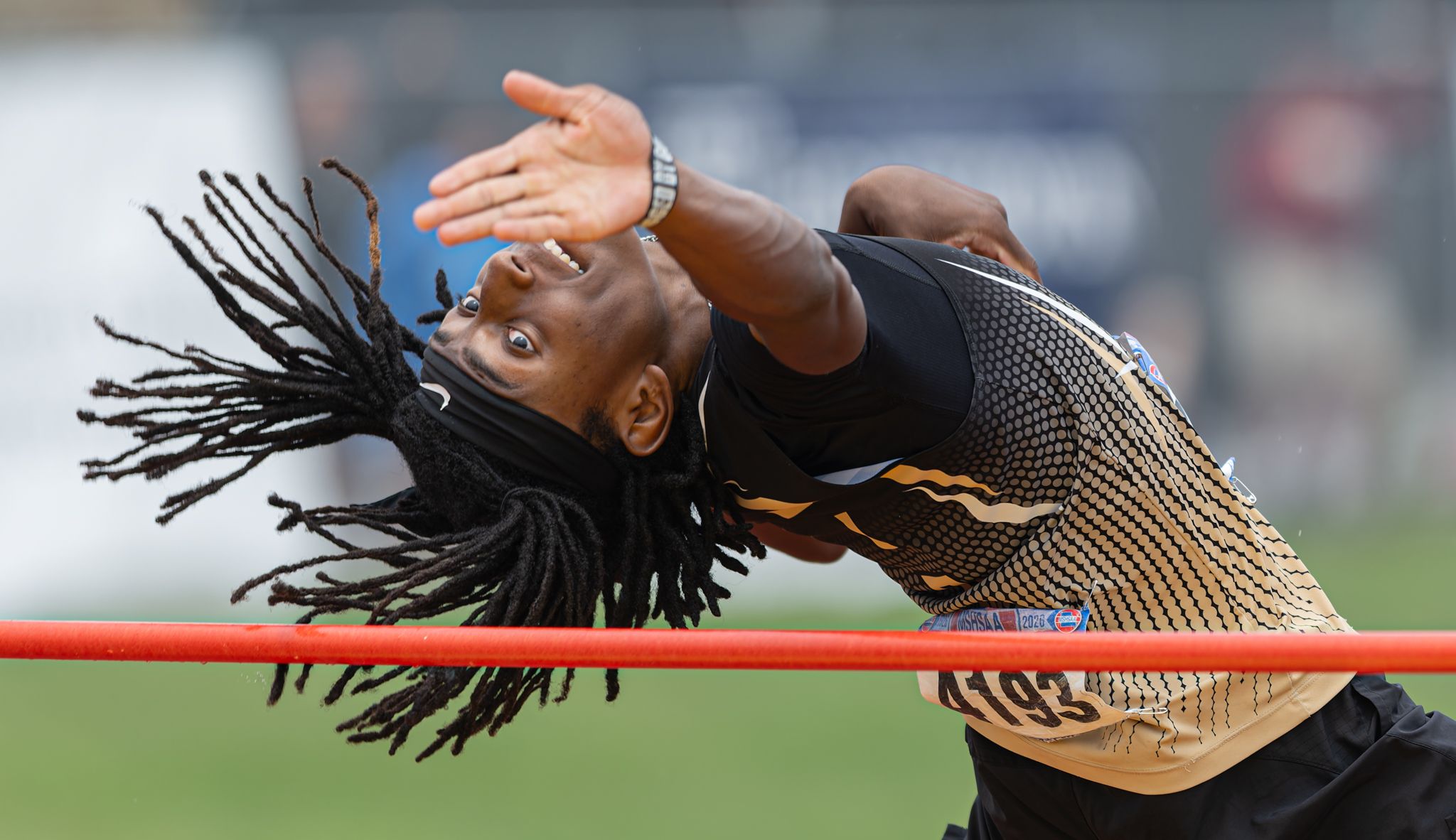 Athlete with dreadlocks performing high jump, arching back over bar in black and gold uniform.