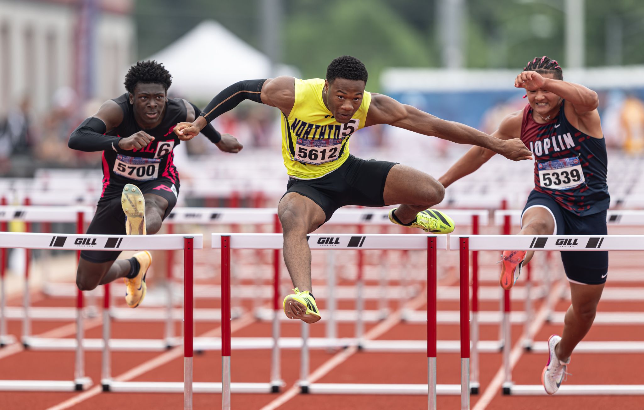 Three athletes compete in a hurdles race at a track and field event, mid-leap over barriers in athletic uniforms.