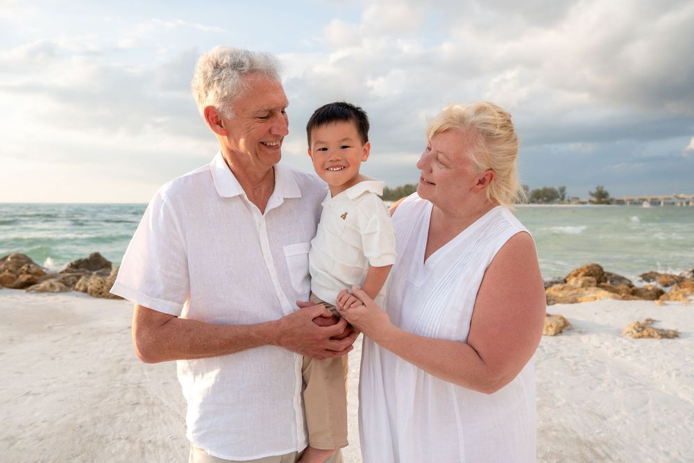 Grandparents smiling and holding their young grandson on a rocky beach with ocean waves in the background.