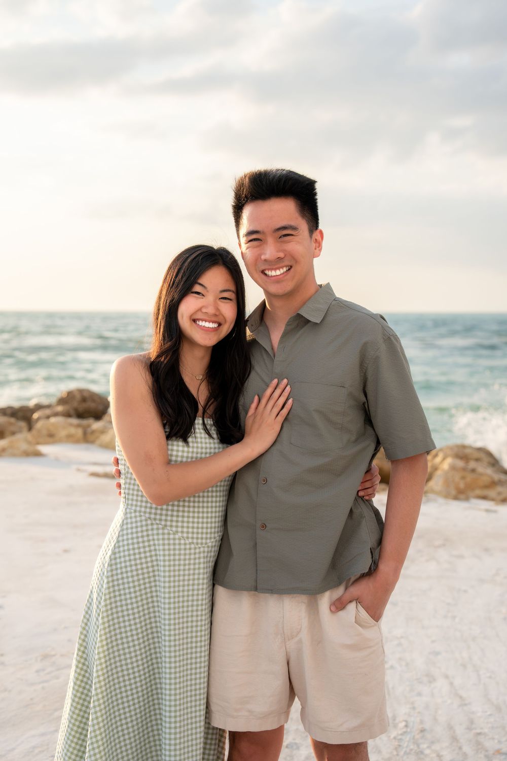 Happy couple smiling together on a beach, woman in green dress, man in olive shirt and khaki shorts.