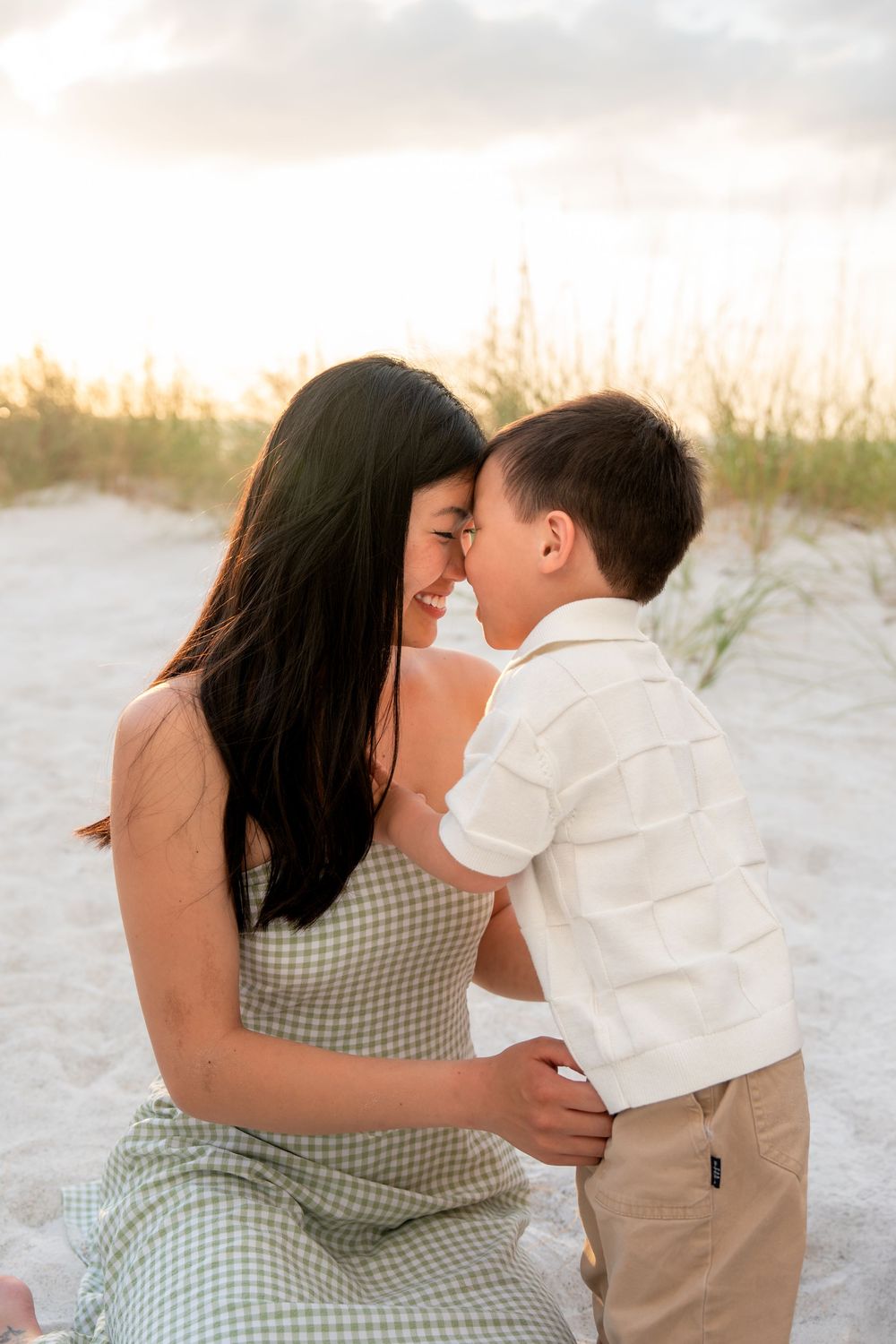 A mother and young son share a tender nose-to-nose moment on a sandy beach at sunset.