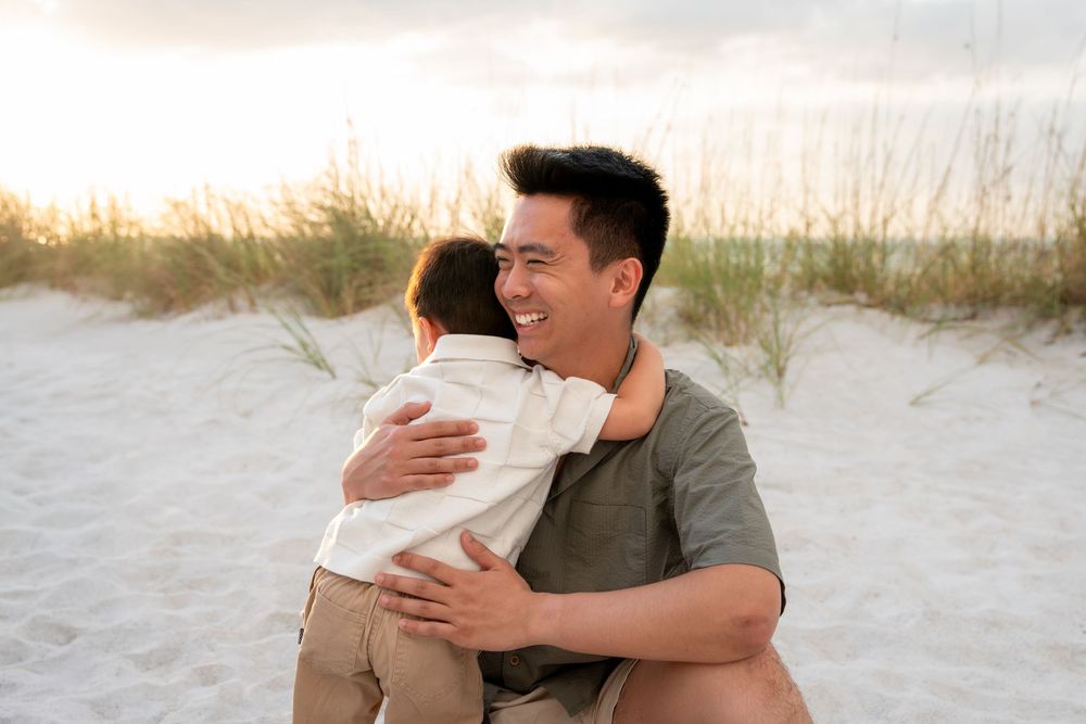 Father hugging young child on sandy beach at golden hour, smiling warmly near sand dunes.