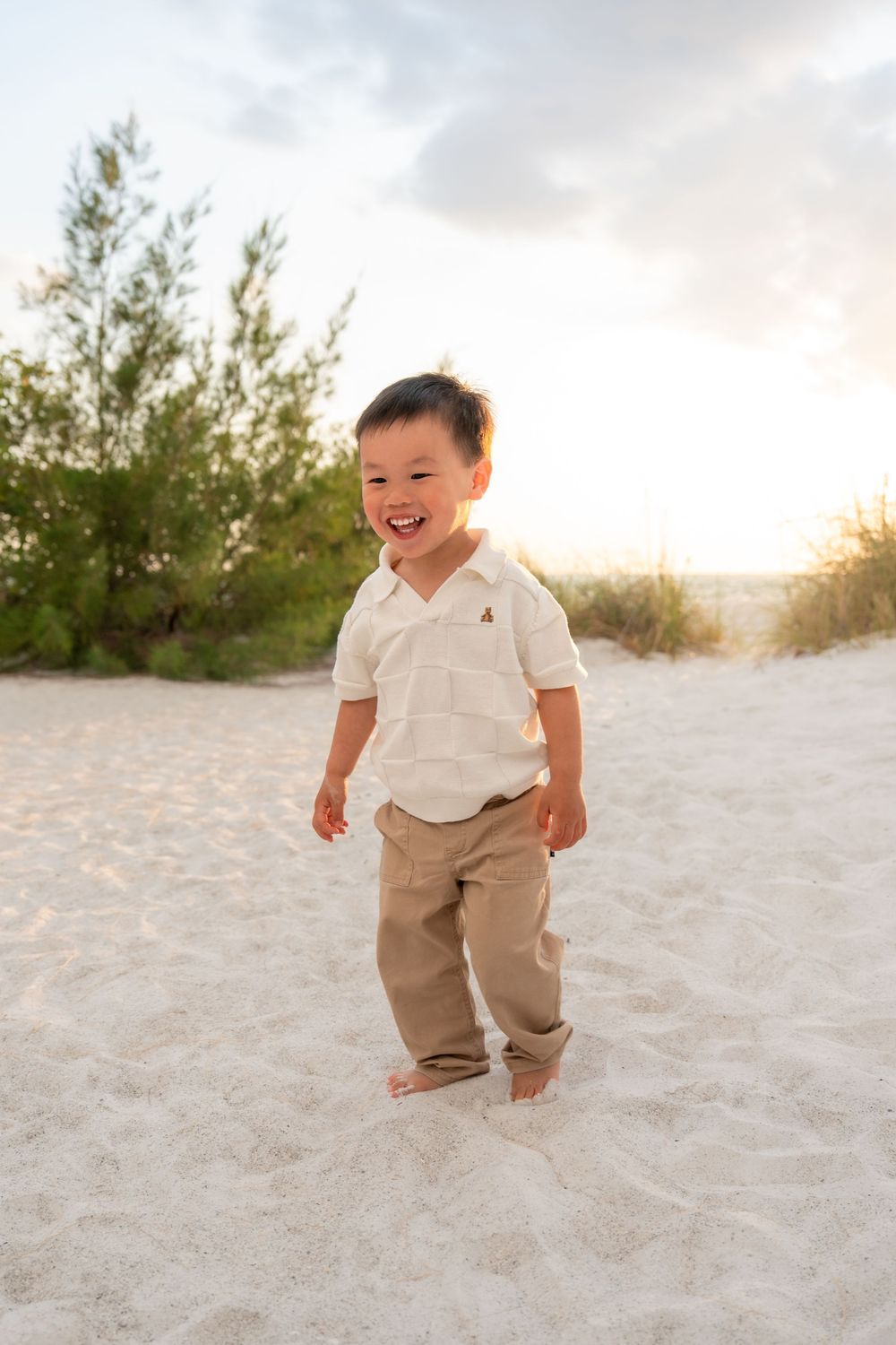 A happy young Asian boy laughs and plays barefoot on white sand beach at sunset.